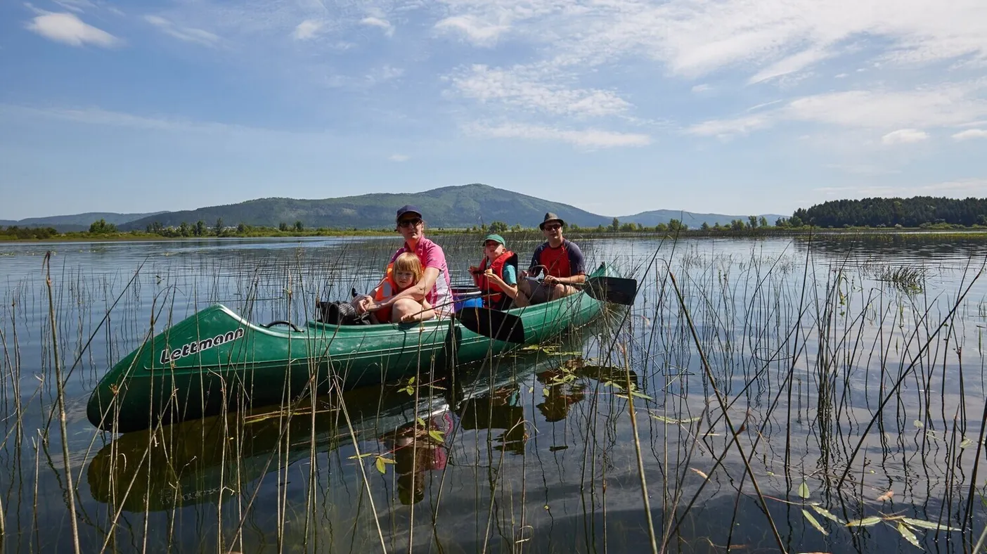 Skupina treh oseb na večjem zelenem kanuju vesla po Cerkniškem jezeru, daleč v ozadju je vidna gora Slivnica.