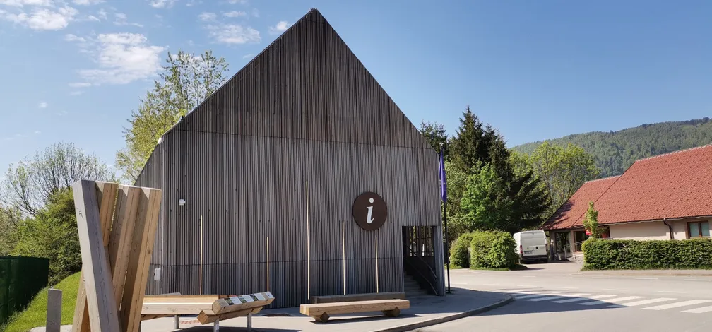 Lesena stavba s strmo, trikotno streho in okroglim znakom z belo črko 'i' na pročelju, ki označuje informacijski center- Center za obiskovalce Cerkniško jezero. V ozadju so drevesa in druge stavbe z rdečimi strehami pod modrim, jasnim nebom.