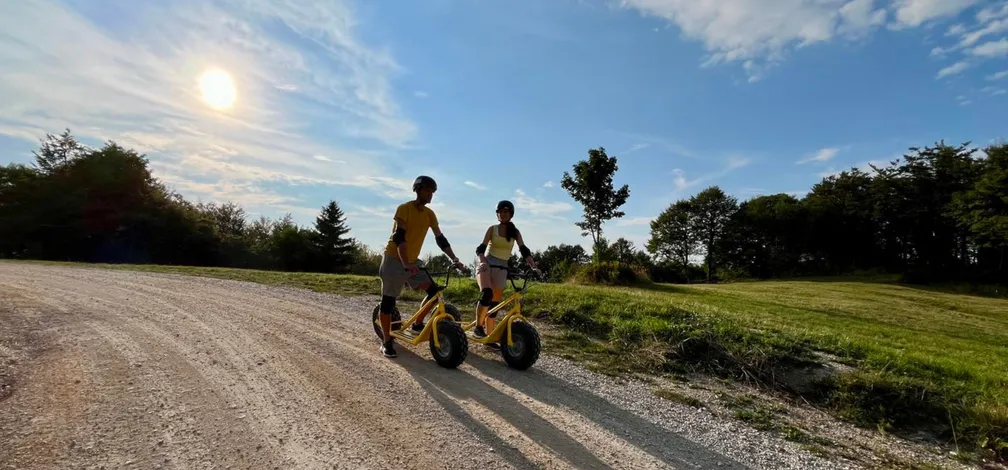Dve osebi se vozita z rumenima skirojema po makadamski poti. Sonce močno sije in meče dolge sence na tla. V ozadju so drevesa in zelenje, kar kaže na naravno okolje.