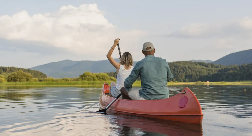 Dve osebi veslata v rdečem kanuju po mirnem jezeru, obdanim z gorami in drevesi. Nebo je delno oblačno.