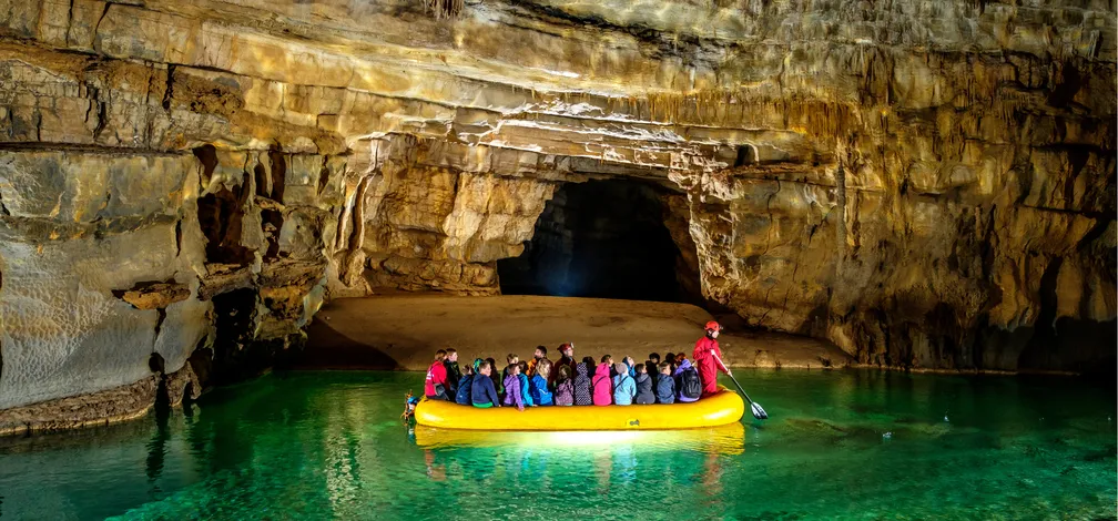 Skupina ljudi v rumenem napihljivem čolnu v veliki jami z osvetljenimi stenami in prozorno zeleno vodo. Jama ima visok strop s skalnimi formacijami, čoln pa vodi oseba v rdečih oblačilih.