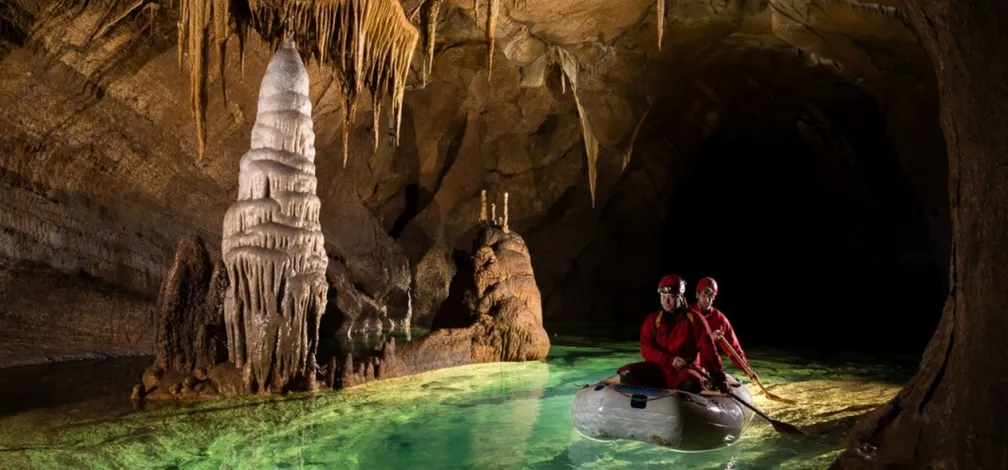 Dve osebi v rdečih oblačilih in čeladah sedita v majhnem napihljivem čolnu v jami. Jama ima kapnike, ki visijo s stropa, in velik stalagmit ob robu vode. Voda je prozorna z zelenkastim odtenkom in odseva jamske formacije. Ozadje jame je temno, kar poudari osvetljen sprednji del prizora.