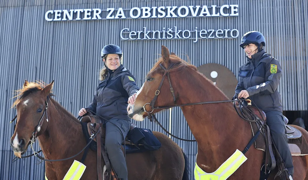 Dva naravovarstvena nadzornika na konjih stojita pred stavbo z napisom “Center za obiskovalce Cerkniško jezero”. Naravovarstvena nadzornika sta v zaščitnih čeladah in uniformah, konja pa sta opremljena z odsevnimi trakovi.