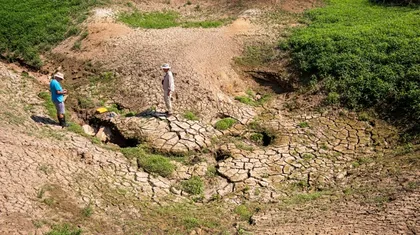 Dve osebi stojita na suhem, razpokanem zemljišču Cerkniškega jezera z redkimi zaplatami zelene vegetacije. Ena oseba nosi modro majico in klobuk, druga pa svetla oblačila in prav tako klobuk. V ospredju je poudarjena suha, razpokana zemlja, v ozadju pa nekaj zelenja.