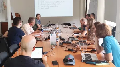 Skupina ljudi sedi okoli konferenčne mize v sejni sobi. Na mizi so prenosniki, tablice in druge elektronske naprave. Na projekcijskem platnu spredaj je prikazano besedilo.