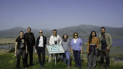 Skupina osmih ljudi stoji na prostem pred informacijsko tablo, v ozadju pa je slikovita pokrajina z gorami in vodnim telesom.